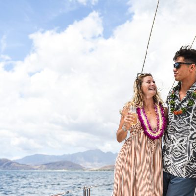 Couple with leis enjoying drinks on a boat with ocean and mountains in the background.