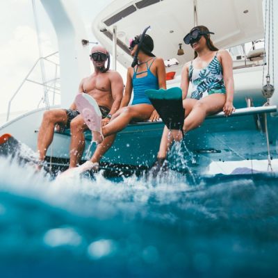 Three people in snorkel gear sit on a boat's edge above clear blue water.
