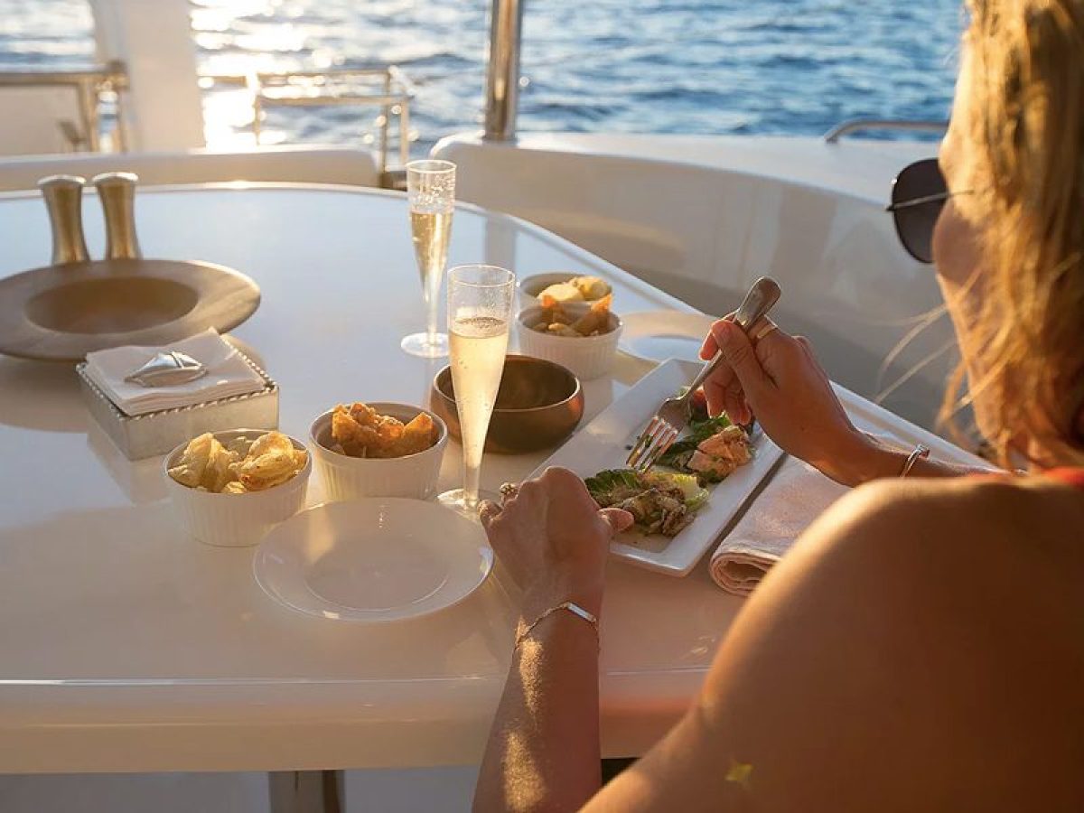 Dining aboard a yacht with food and drinks on a table, ocean in background.