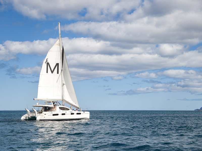 A sailboat with an 'M' on its sail cruising on the sea under a partly cloudy sky.