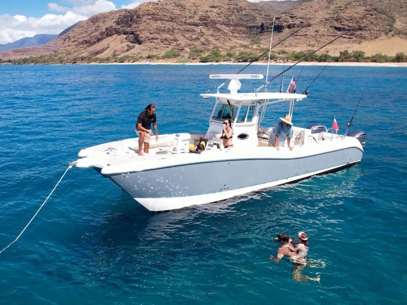 Boat on blue ocean water with people onboard and swimming nearby, mountains in background.