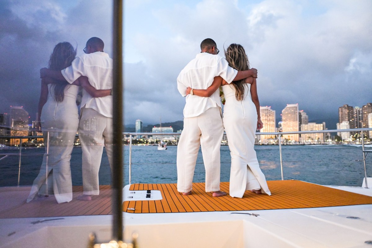 Two people in white outfits embrace on a yacht, city skyline in the background.