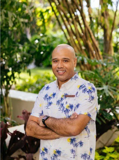 Bald man in floral shirt smiling with arms crossed, standing outdoors in a garden.